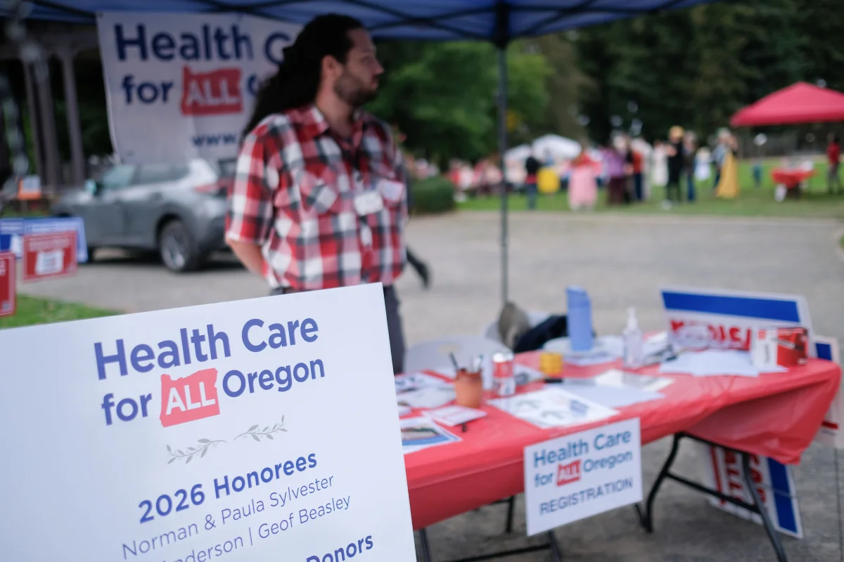 A man stands at a vendor table outdoors. He has signs, pins, and other information about "Health Care for All Oregon."