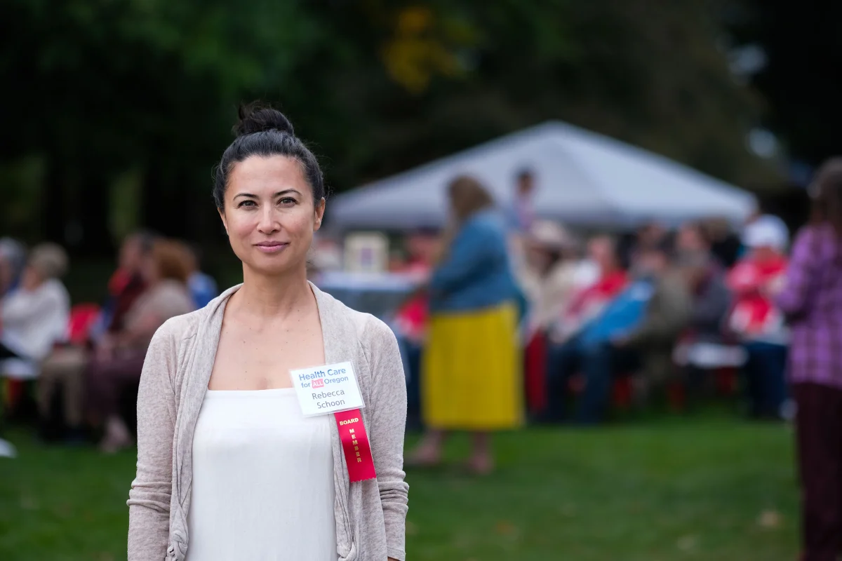 A woman with dark, straight black hair tied up in a bun stands for a photo. She has a plastic name tag attached to her shirt that says, "Healthcare for All Oregon / Rebecca Shcoon."