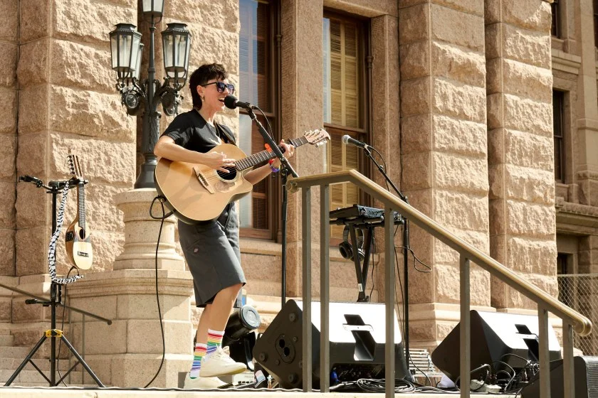 A person in a black T-shirt plays guitar on the steps of a state building on a sunny day.